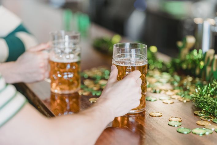 Crop anonymous male friends with glasses of beer sitting at wooden counter while celebrating Saint Patricks day on blurred background